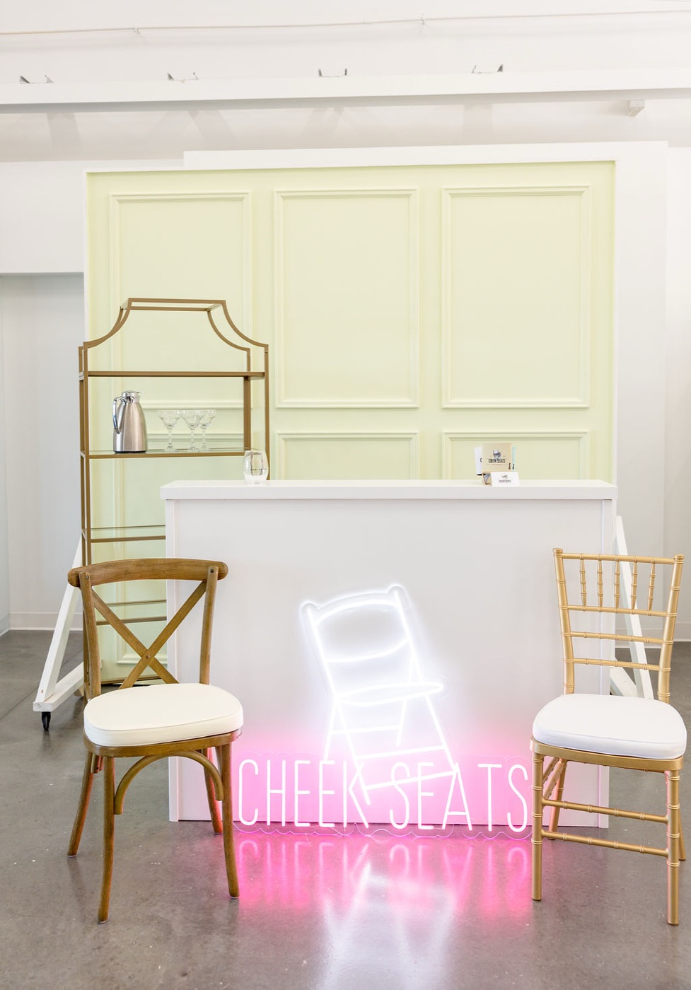 Cheek Seats neon sign with a wood crossback chair, gold chiavari chair, and gold bar shelf